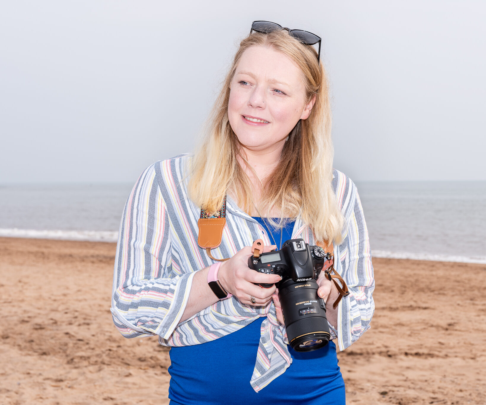 Gemma Varney Photography and Business Mentor Gemma Varney Holding her camera on the beach with a smile, sunglasses on head and blue dress, the light is well balanced on her and the background