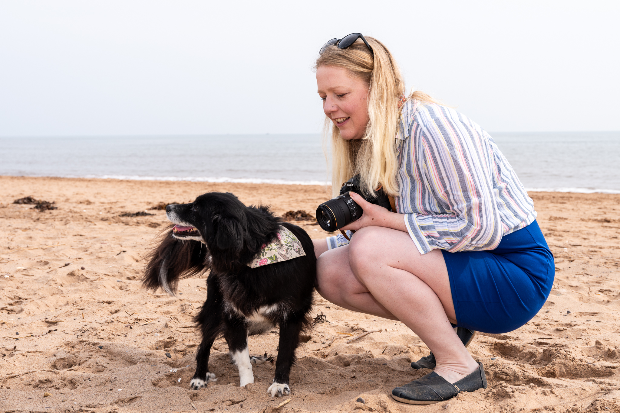 Gemma stroking her collie dog on the beach whilst holding her camera with sunnies on her head and a royal blue dress on a cloudy day