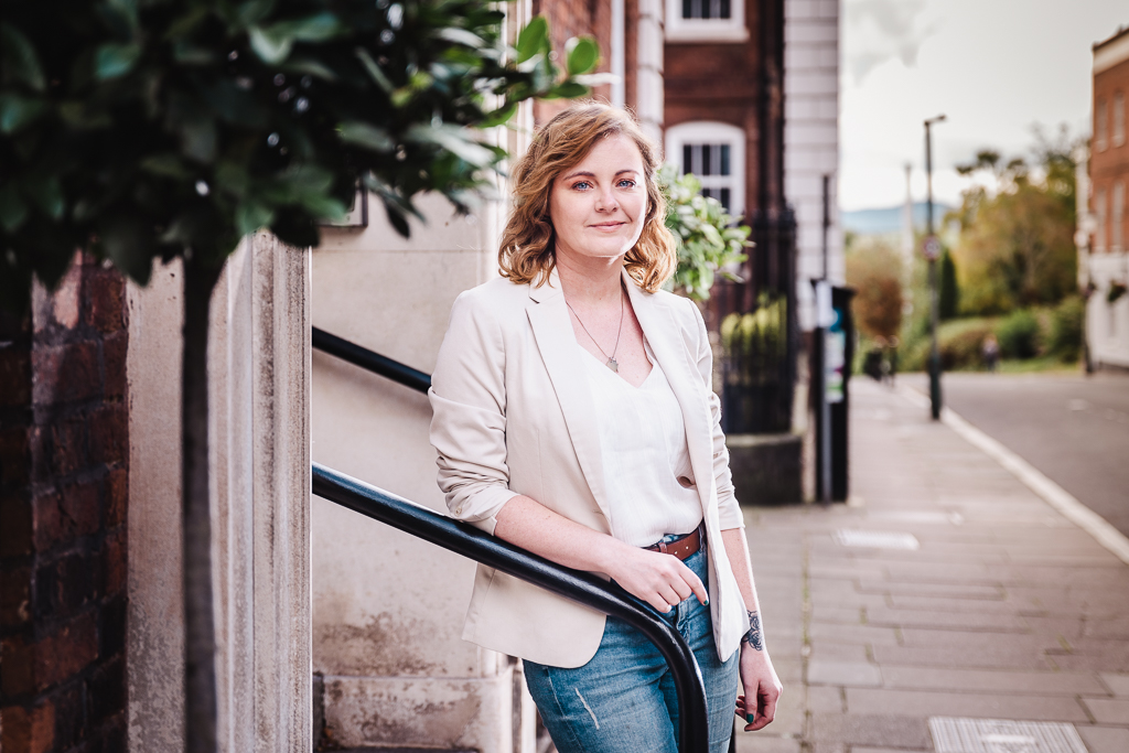 Woman nicely flash lit with soft light leaning on a railing outdoors wearing a cream suit jacket and bright white vest top.